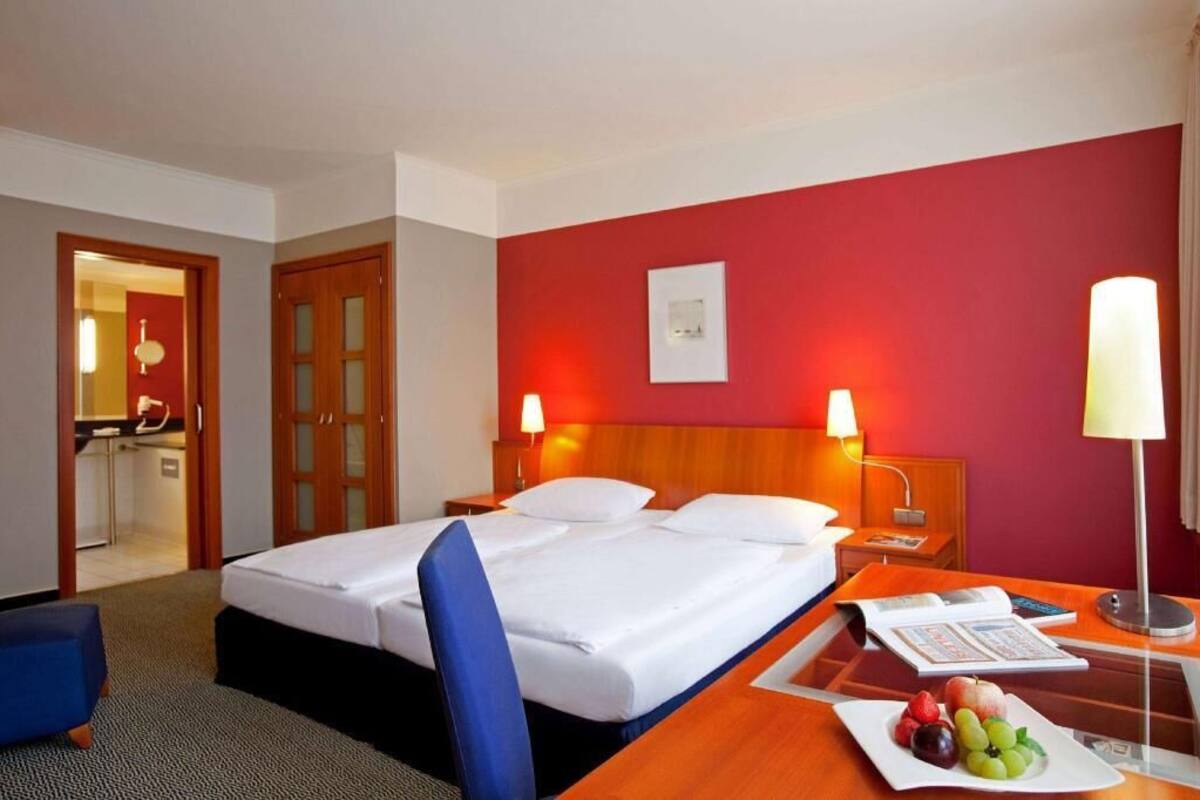 A hotel room with a bed, two bedside lamps, a desk with a fruit plate, and a view of a kitchen area.