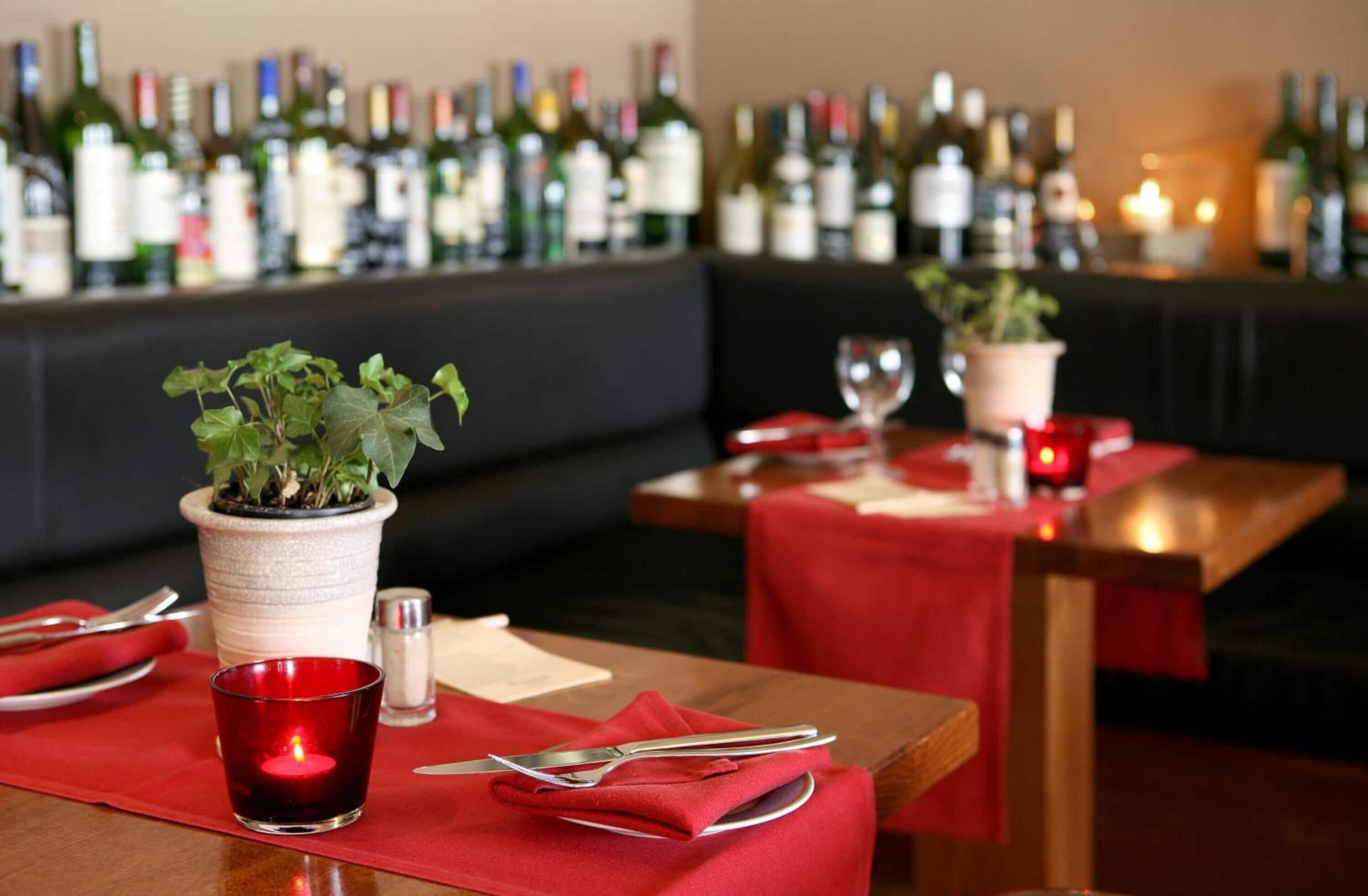 A restaurant setting with tables set for dining, a bar in the background with various bottles, and potted plants on the tables.
