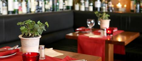 A restaurant setting with tables set for dining, a bar in the background with various bottles, and potted plants on the tables.