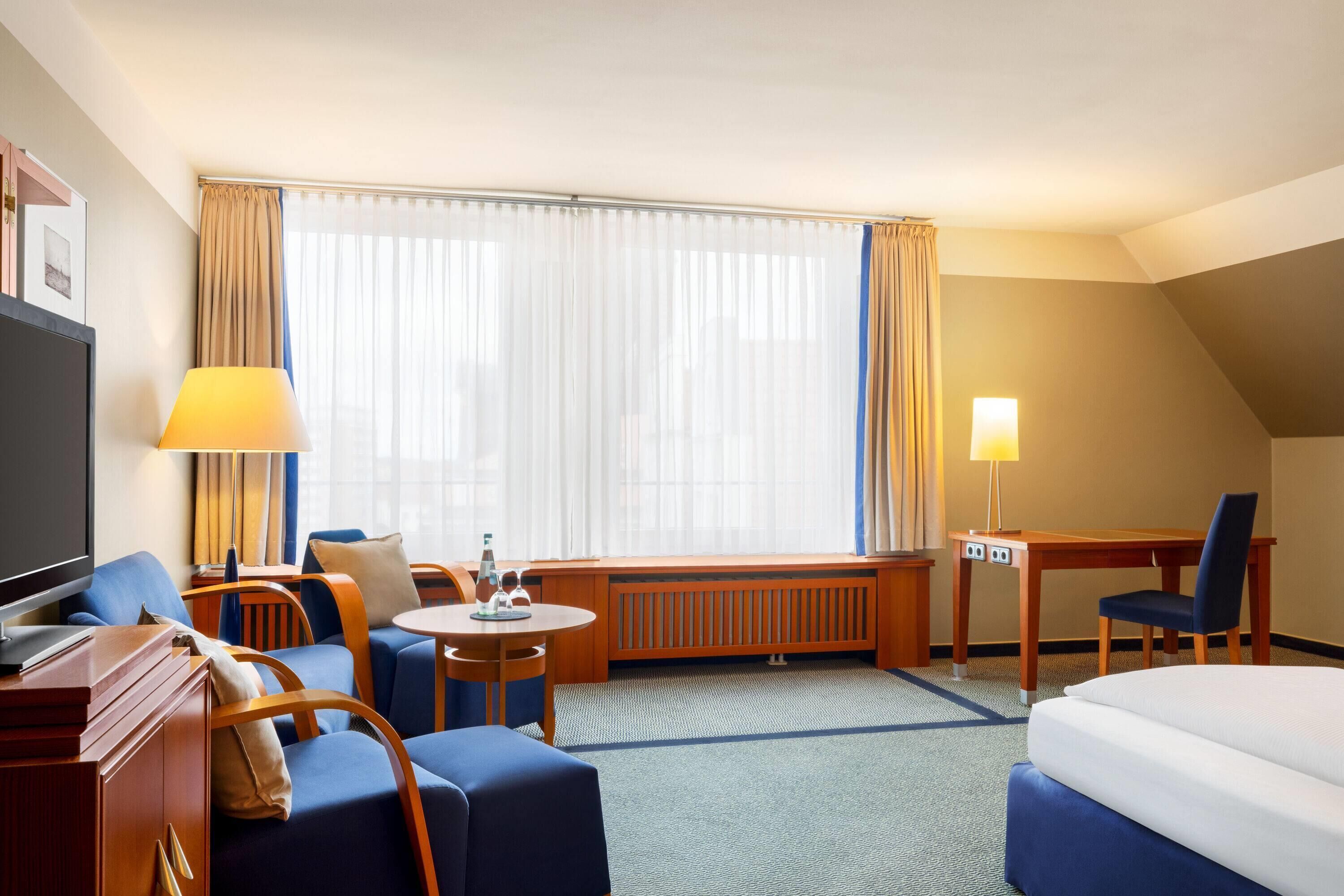 A hotel room with a bed, a desk, a chair, a sofa, a TV, and a window with curtains.