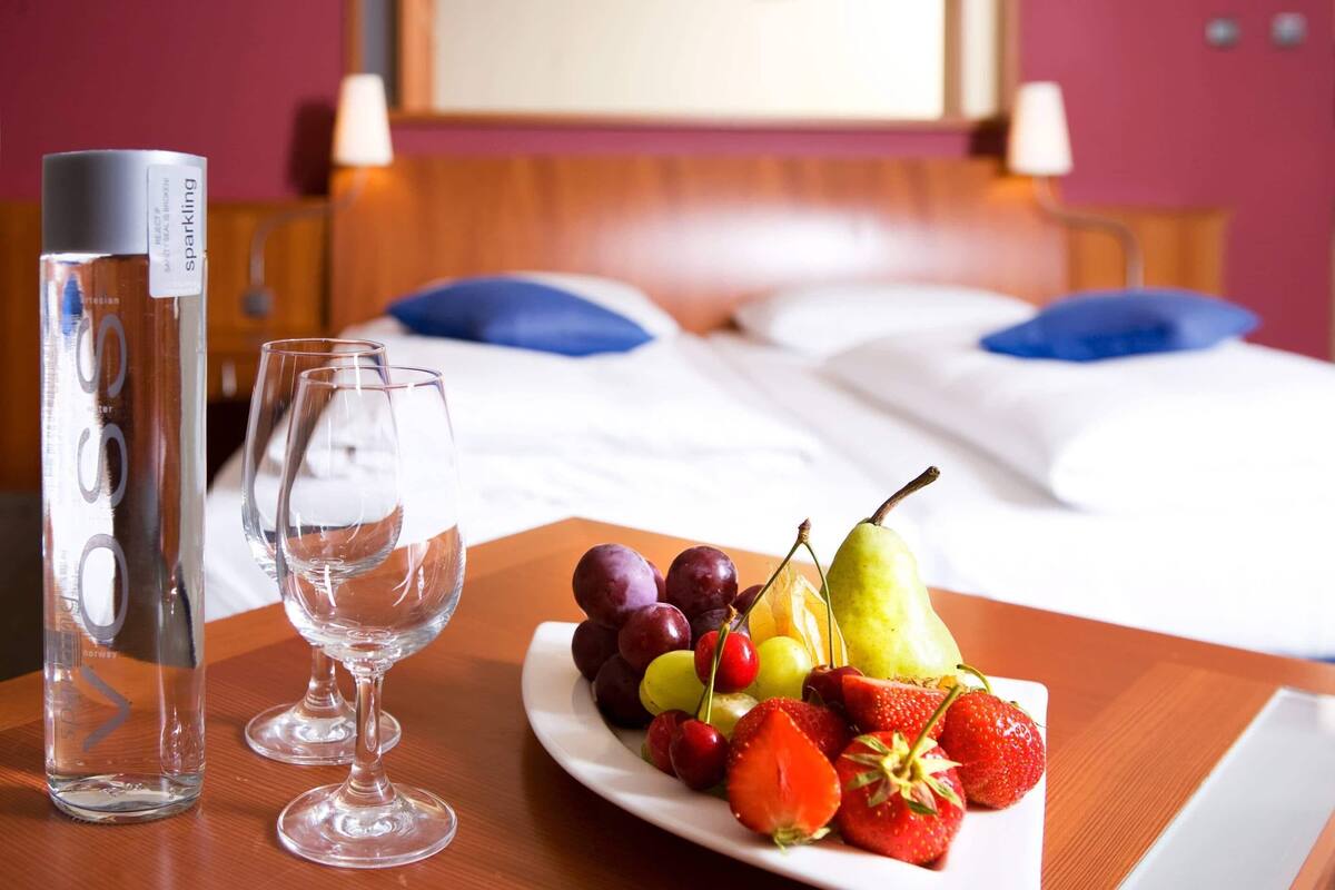 A hotel room with a bed, a bedside table with a bottle and glasses, and a plate of fruit.