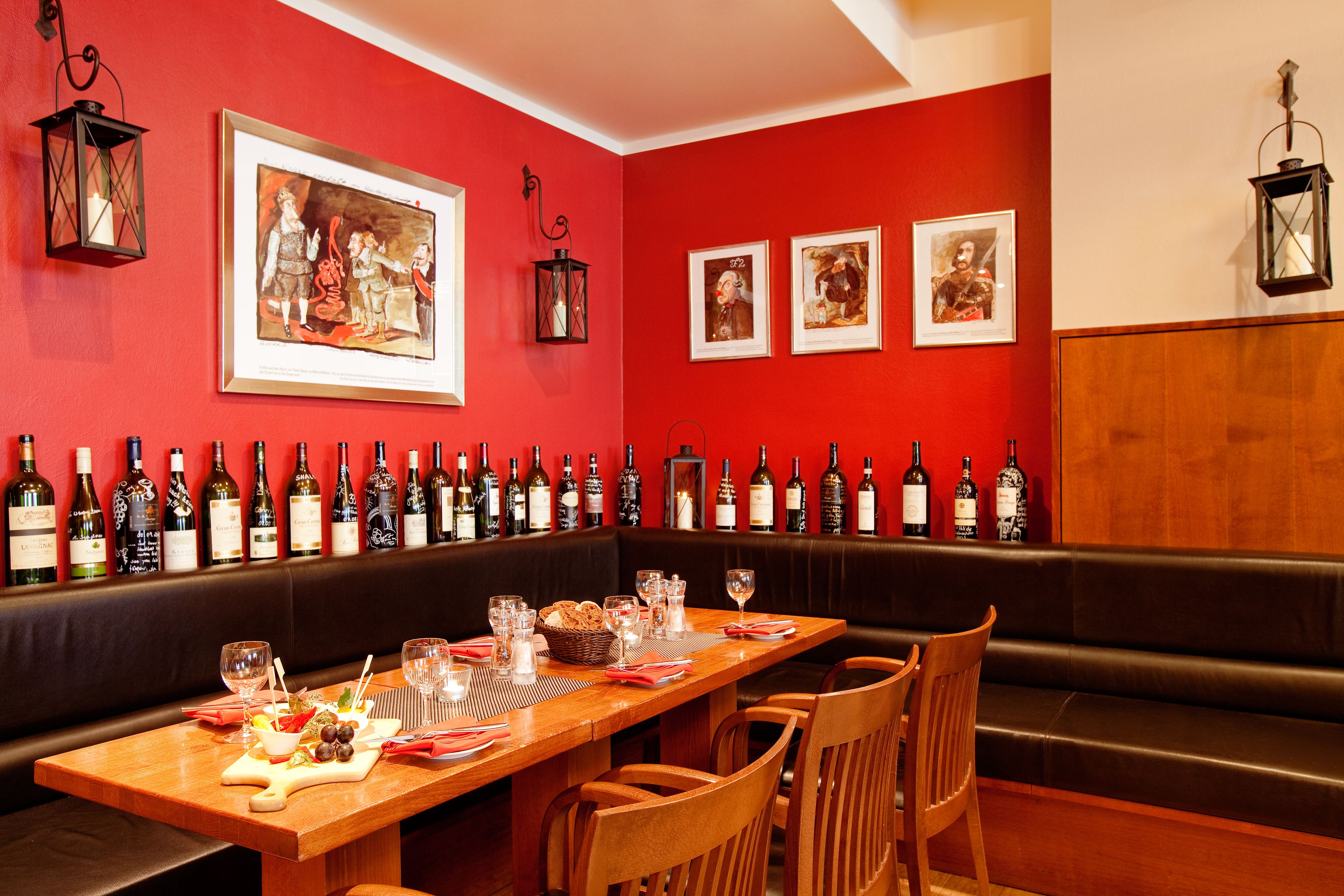 Un restaurant avec une longue table en bois dressée pour le dîner, un mur rouge orné de tableaux encadrés et une rangée de bouteilles de vin alignées sur le mur.