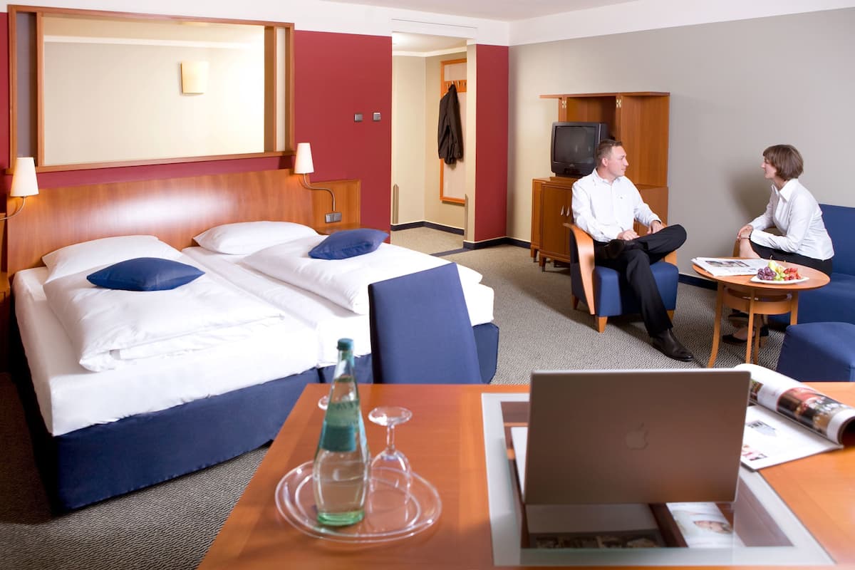 A hotel room with a bed, a desk with a laptop, a TV, and a seating area with two chairs.