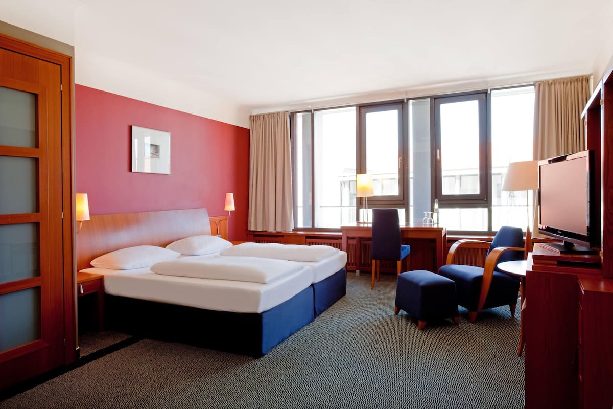 A hotel room with a bed, desk, chair, TV, and large windows.