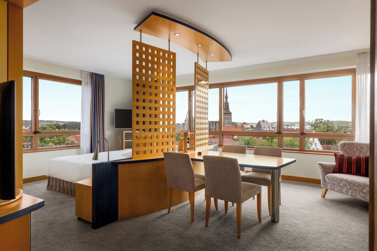 A hotel room with a bed, a desk, a chair, a TV, and large windows offering a city view.