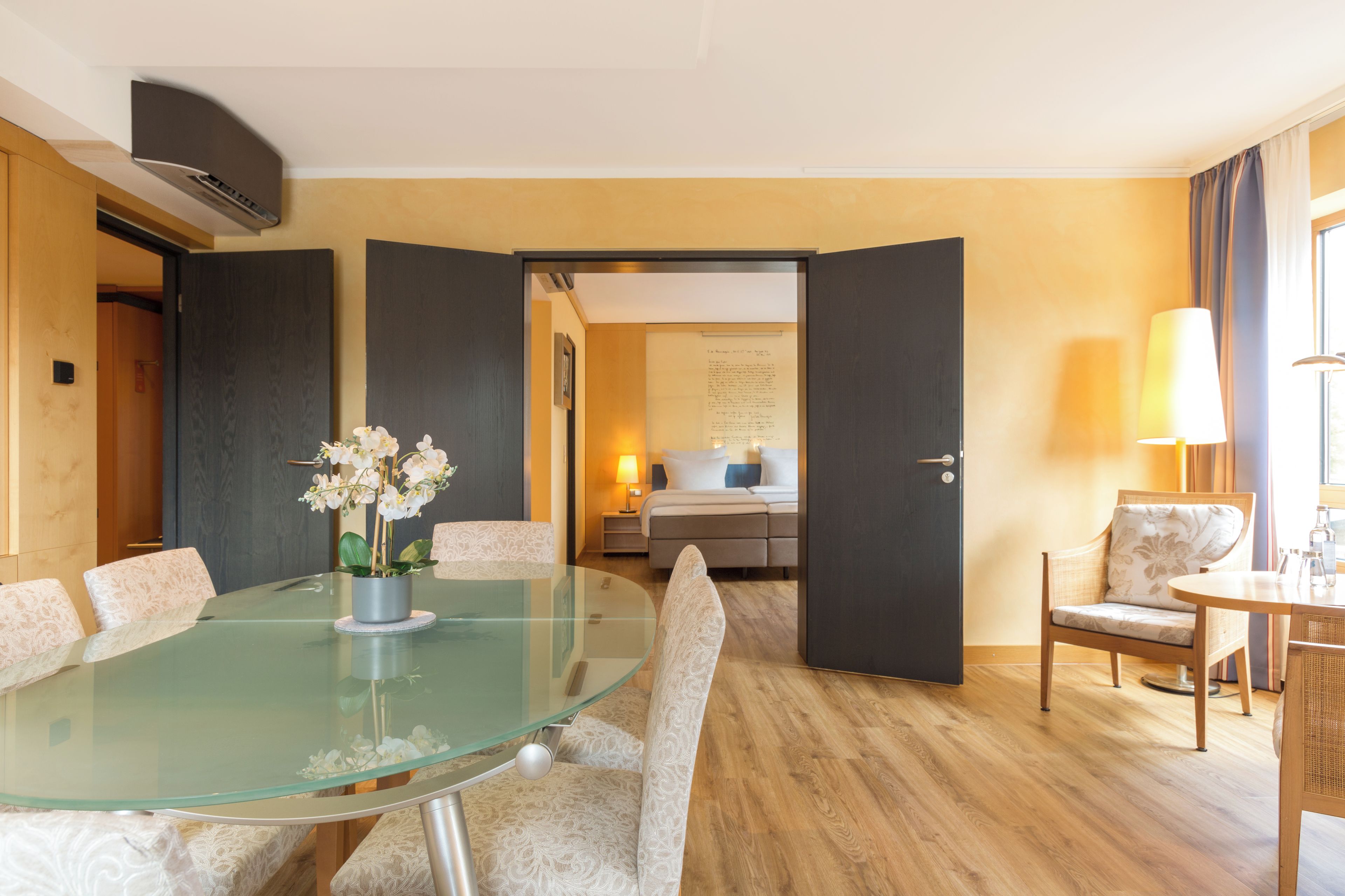 A hotel room with a glass dining table, a bed, a bedside lamp, and a view of the hallway.