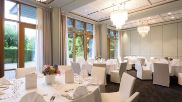 A hotel dining room with multiple tables set for dining, large windows with curtains, and chandeliers.