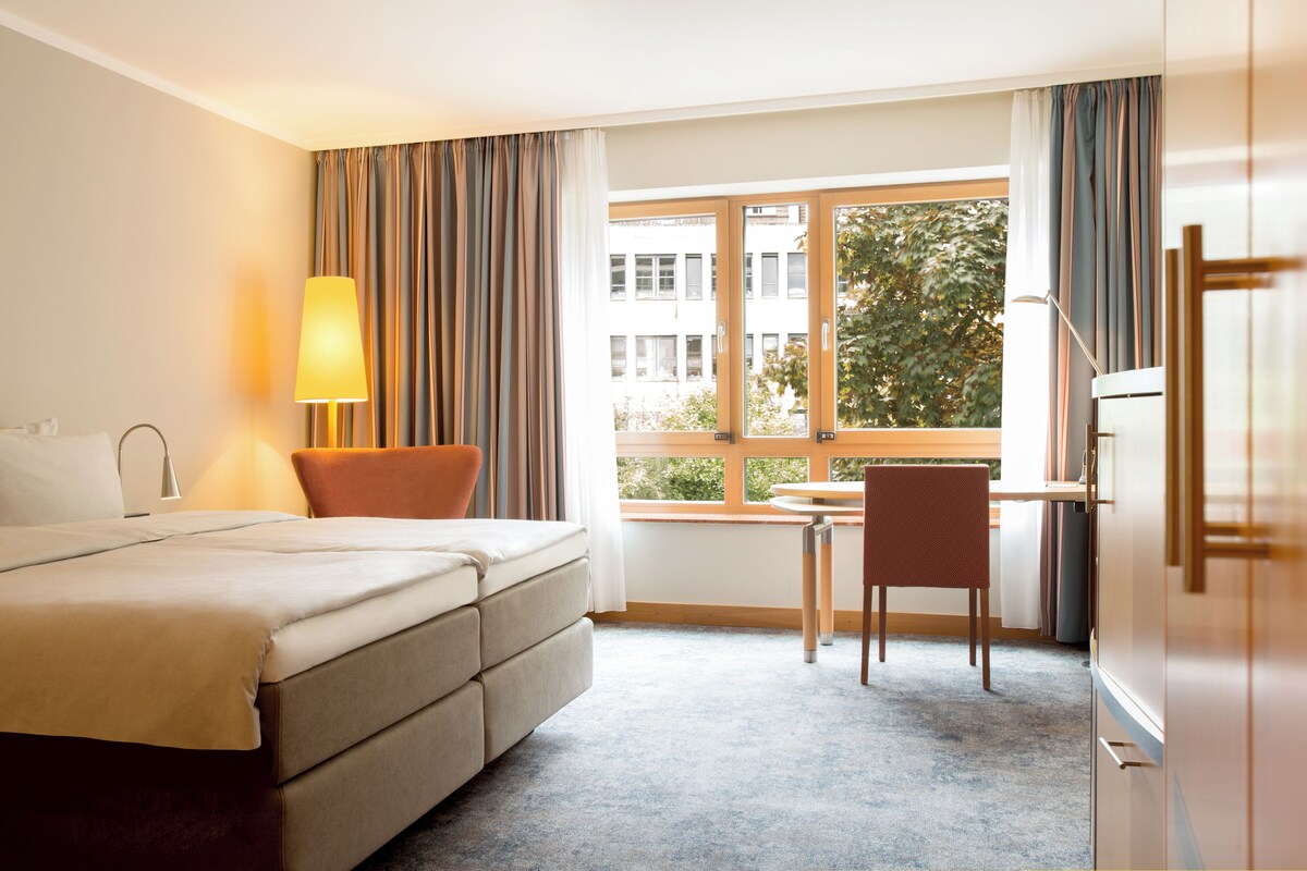 Habitación de hotel con cama, escritorio, silla y ventana con vistas a edificios y árboles.