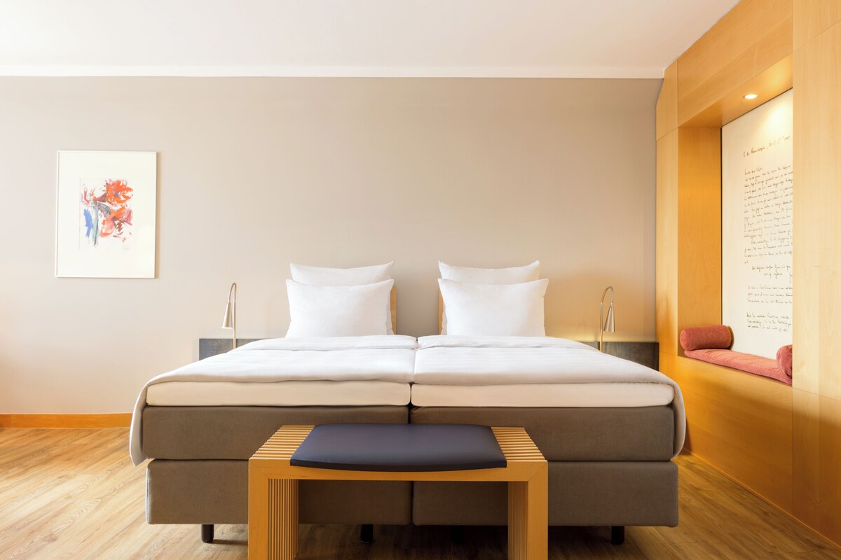 Una habitación de hotel moderna con una cama grande, mesitas de noche, una mesita pequeña y una obra de arte colgada en la pared.