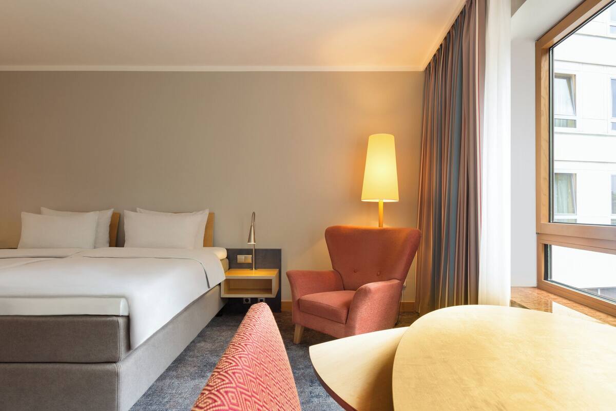 Habitación de hotel con cama, mesita de noche, lámpara, silla y vista al exterior.