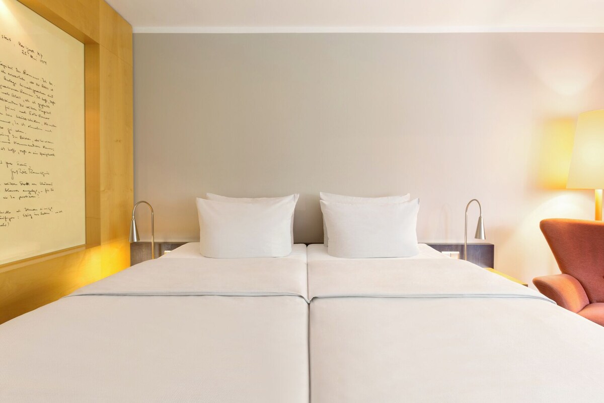 Una cama doble con sábanas blancas, cabecera de madera, mesita de noche con lámpara y una silla.