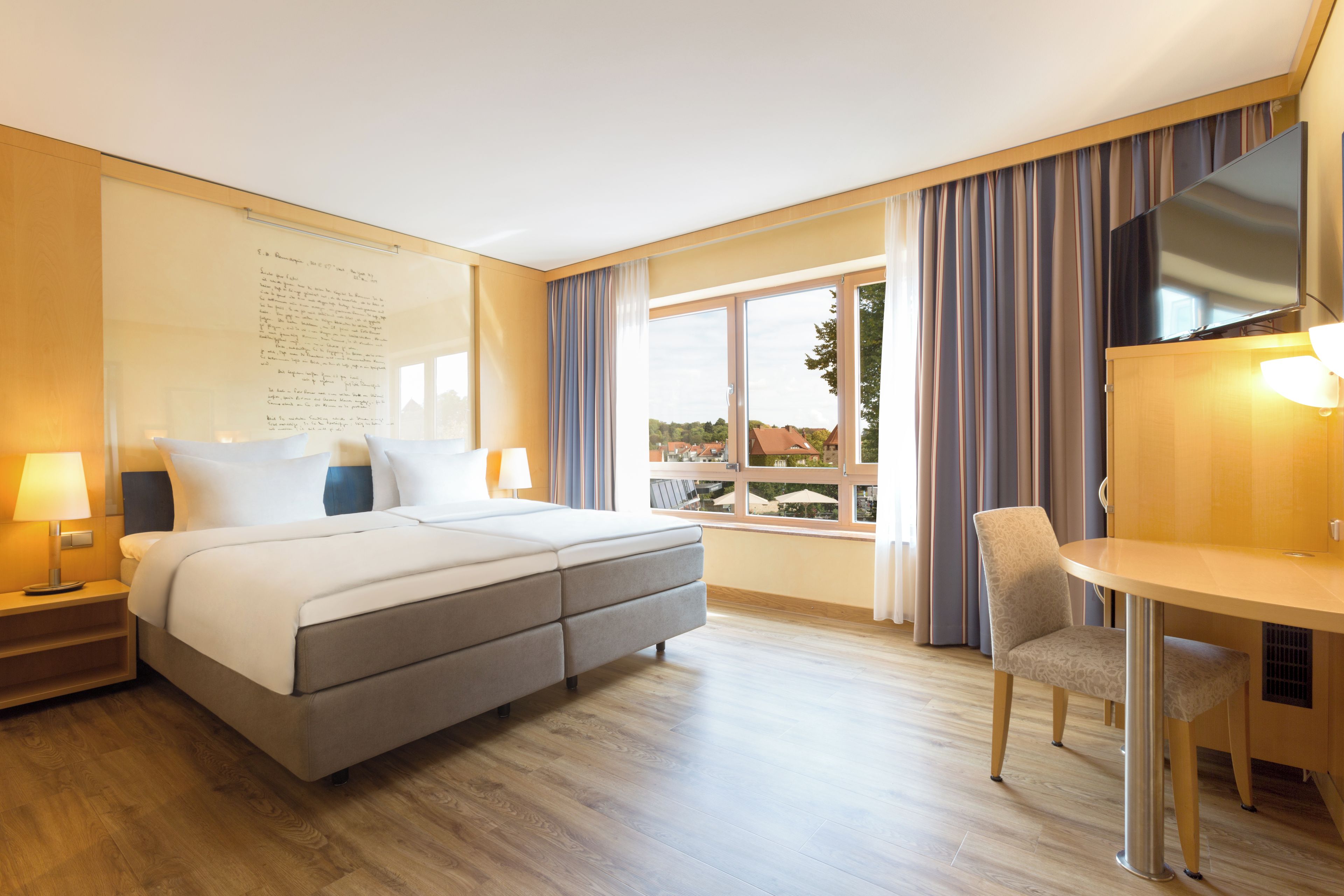 A hotel room with a large bed, a desk, and a chair. There is a window with curtains and a view of buildings outside.