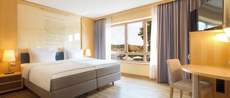 A hotel room with a large bed, a desk, and a chair. There is a window with curtains and a view of buildings outside.