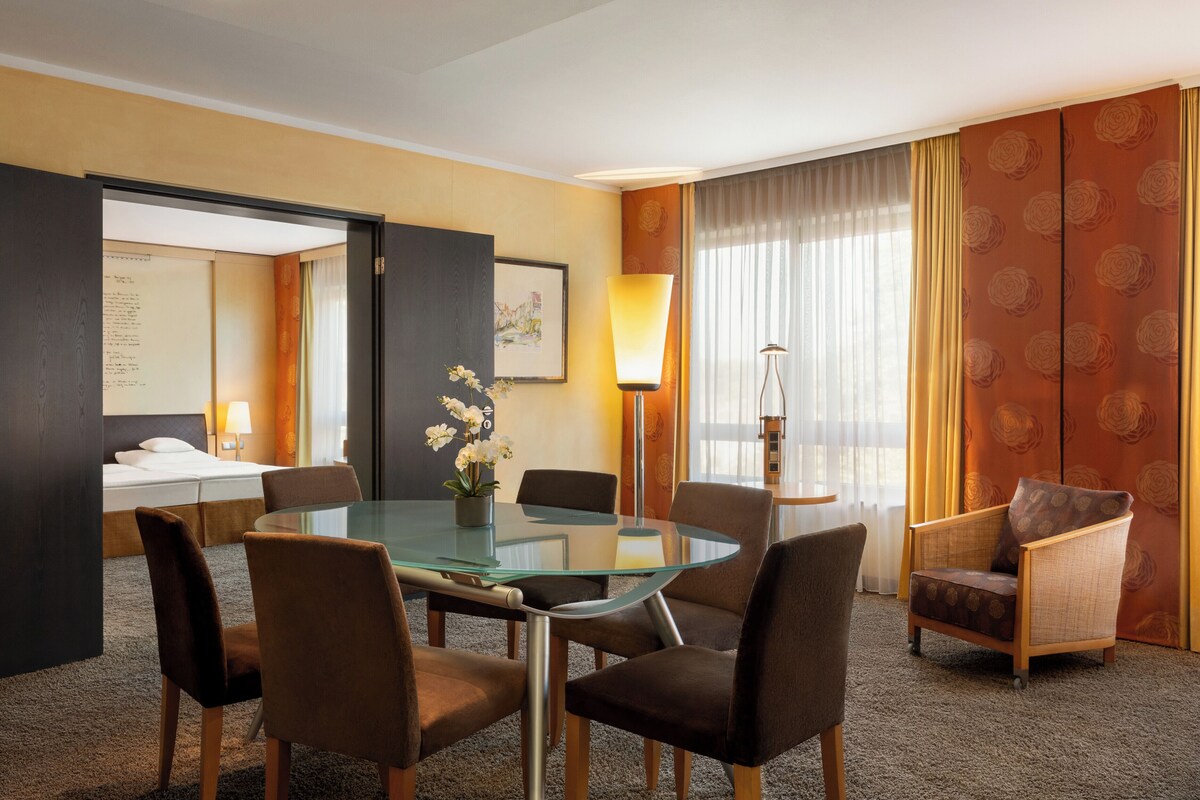 A hotel room with a glass table, four chairs, a sofa, a bed, a desk with a lamp, a plant, and a large window with curtains.