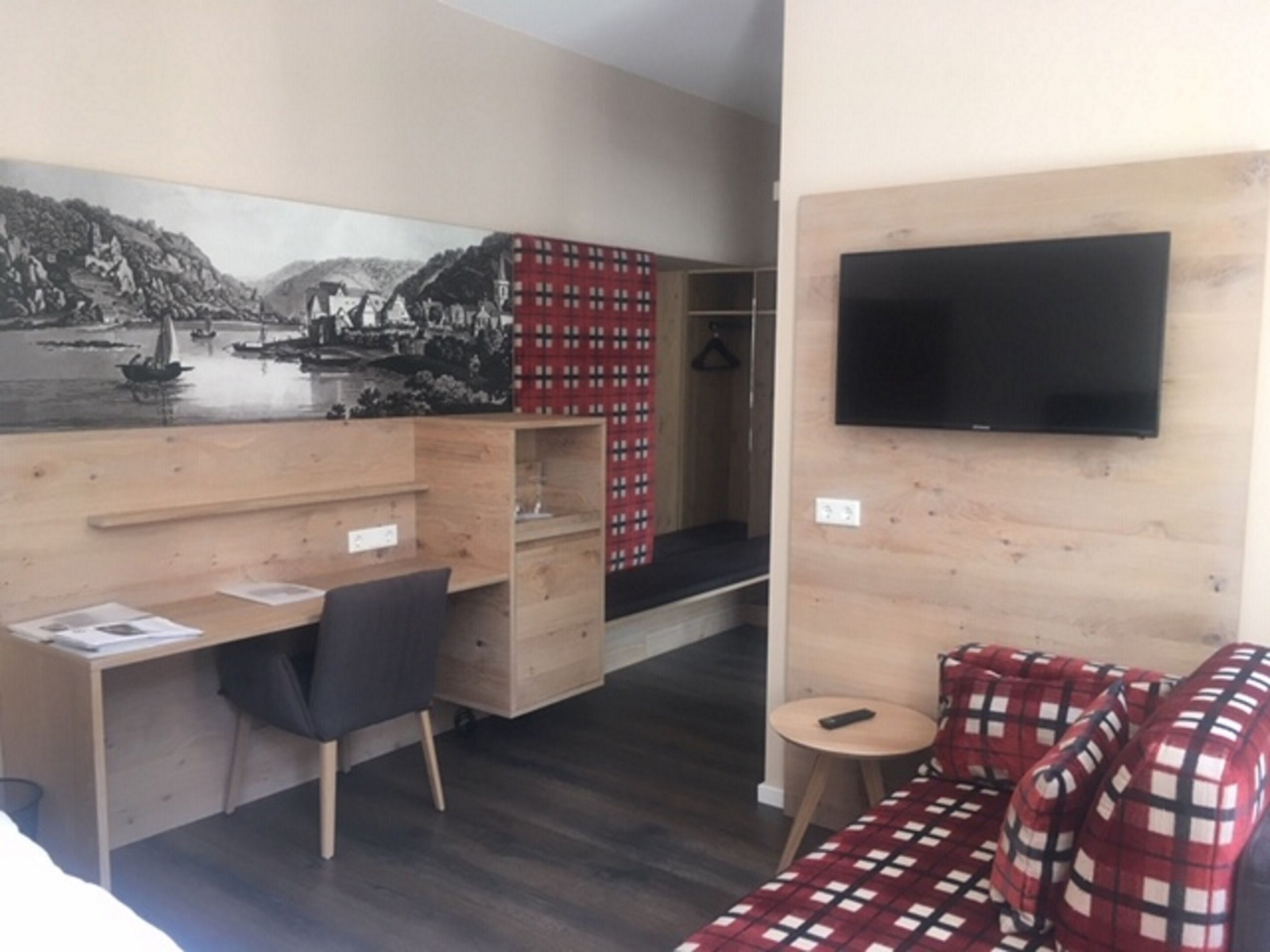 A room with a wooden desk, a chair, a red plaid sofa, a television mounted on the wall, and a large landscape painting.