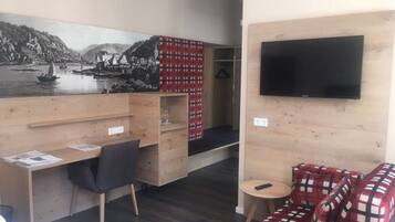 A room with a wooden desk, a chair, a red plaid sofa, a television mounted on the wall, and a large landscape painting.
