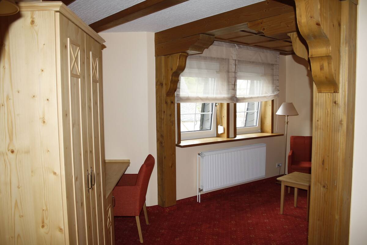 Una habitación con un armario de madera, alfombra roja, ventana con cortinas translúcidas, radiador y una mesa con una silla.