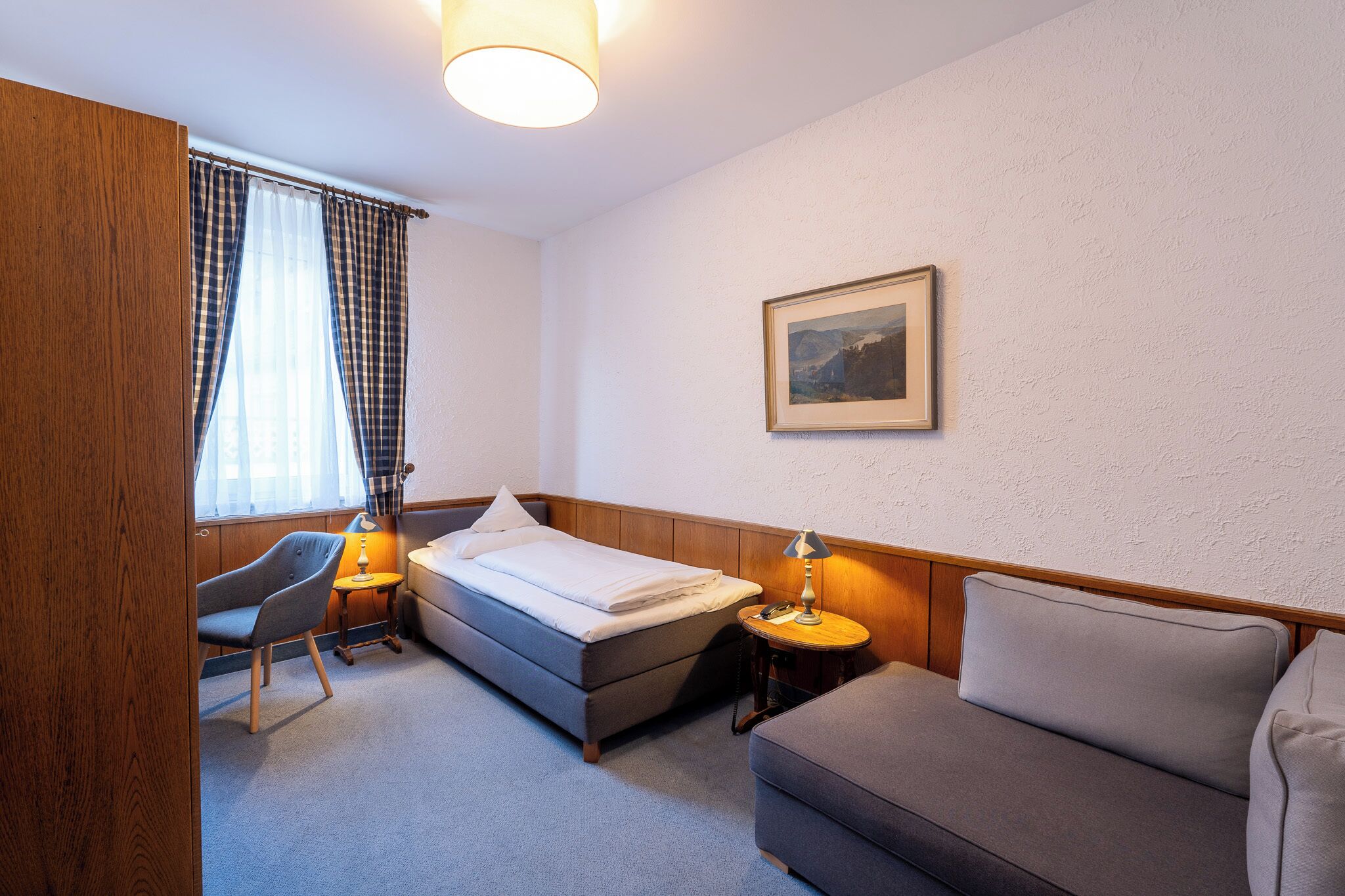 Ett hotellrum med en säng, en soffa, en stol, en lampa, en tavla på väggen och ett fönster med gardiner.
