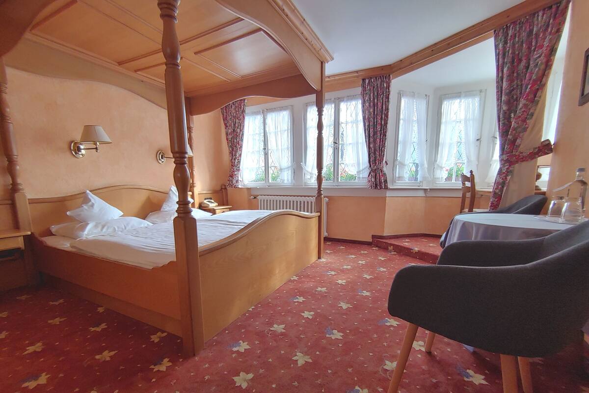Una cama con dosel y cabecero de madera, una mesita de noche con lámpara, una ventana con cortinas y una pequeña mesa con sillas en una habitación de hotel.