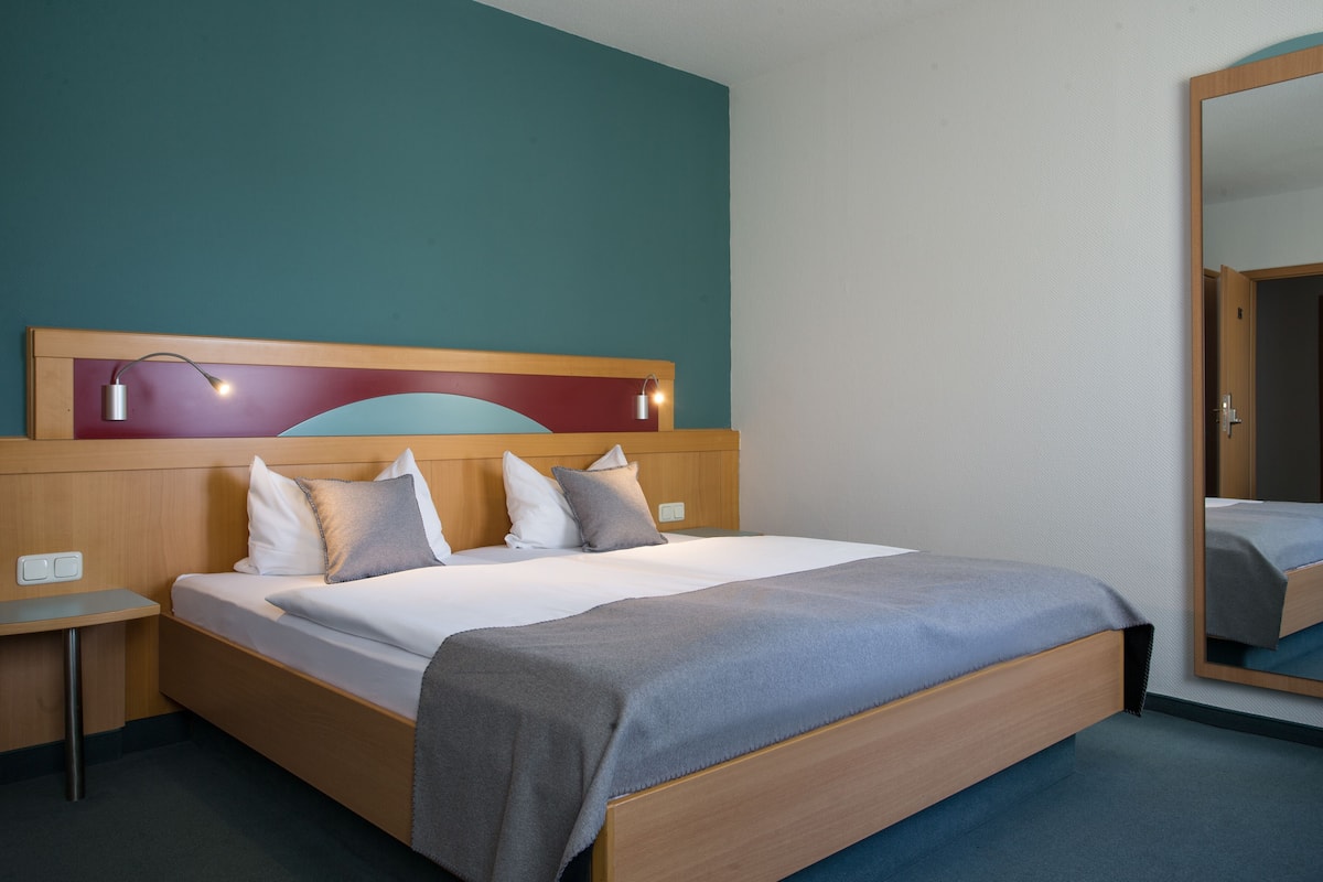 A hotel room with a wooden bed, a nightstand, a mirror, and a teal-colored wall.