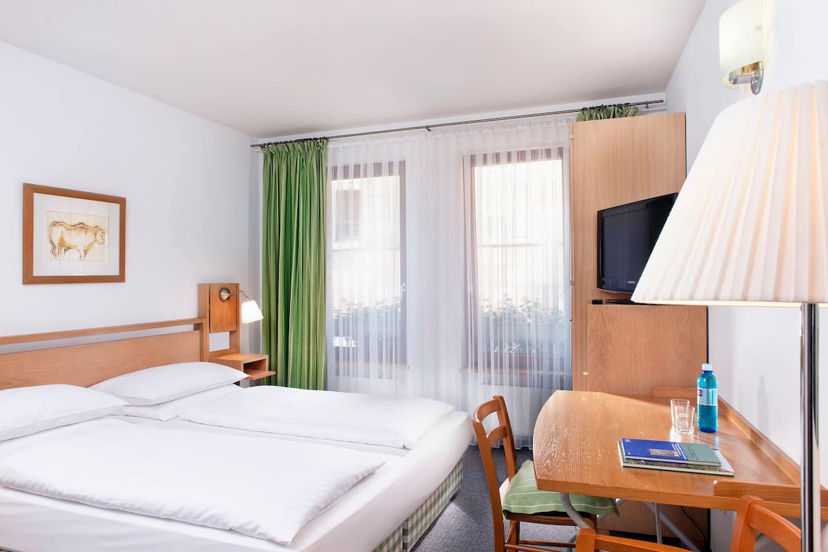 A hotel room with a bed, a desk, a chair, a lamp, a TV, and a window with curtains.