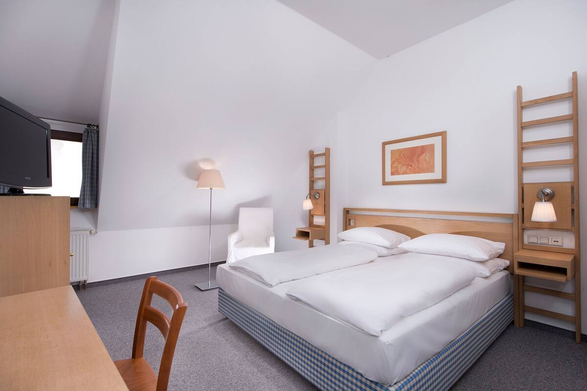 A hotel room with a bed, a desk, a chair, a floor lamp, a ladder, and a framed picture on the wall.