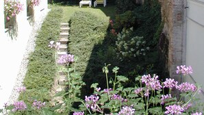 Um pátio estreito com muro de tijolos, um edifício branco com janelas e um jardim com flores e um caminho.