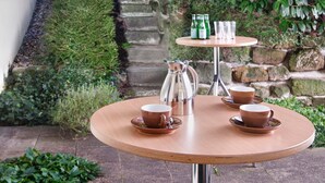 A round wooden table with four cups and saucers, a coffee pot, and two bottles of beer, set against a backdrop of lush greenery and stone steps. - Hotel Agneshof Nürnberg (Nuremberg)