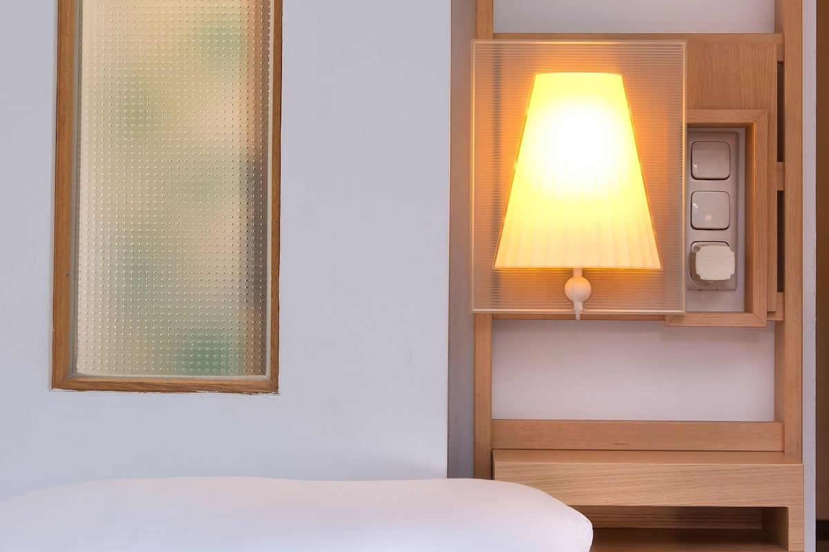 A hotel room with a bed, a wall-mounted lamp, and a frosted glass window.