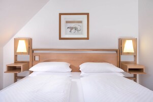 Una cama doble con ropa de cama blanca, cabecera de madera, dos mesitas de noche con lámparas y un cuadro en la pared.