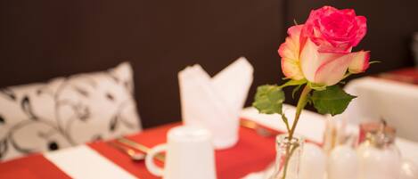 A dining table with a red tablecloth, white napkins, a white pitcher, and a vase with a pink rose.