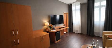Habitación de hotel con un armario de madera grande, un televisor de pantalla plana, un escritorio con lámpara, una mesita con bandeja y un ventanal con cortinas.
