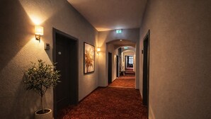 A hotel hallway with a carpeted floor, wall-mounted light fixtures, framed artwork, and an exit sign. - Hotel Bayrischer Hof (Heidelberg)