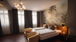 A hotel room with a bed, two armchairs, a small table, a nightstand with a lamp, and a large tropical mural on the wall. - Hotel Bayrischer Hof (Heidelberg)