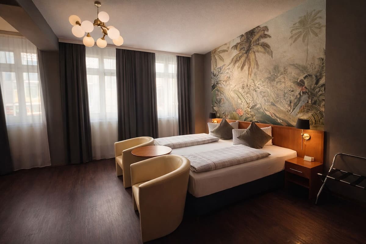 A hotel room with a bed, two armchairs, a small table, a nightstand with a lamp, and a large tropical mural on the wall.