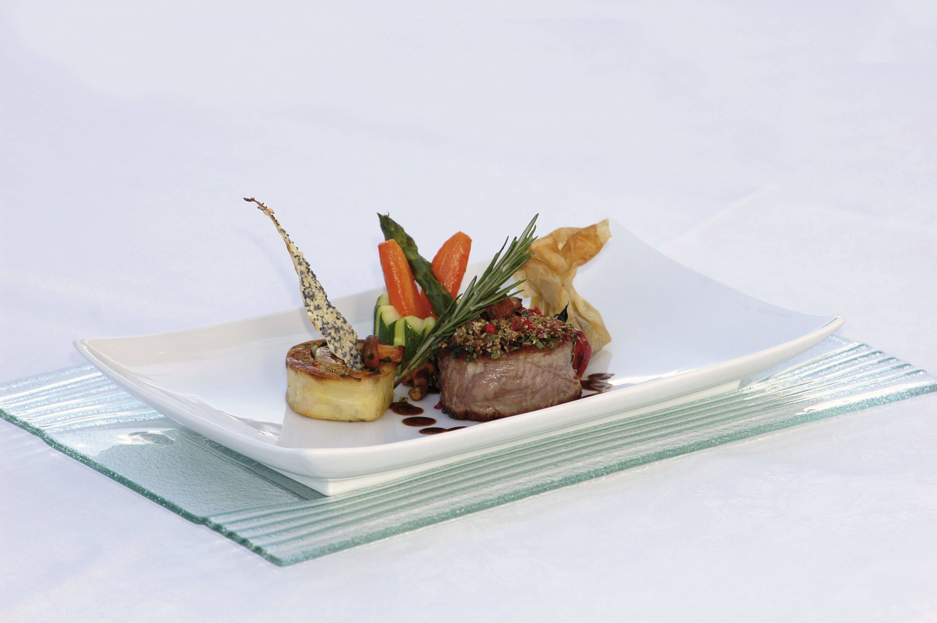 A white plate with a piece of meat, vegetables, and a pastry on a glass table.