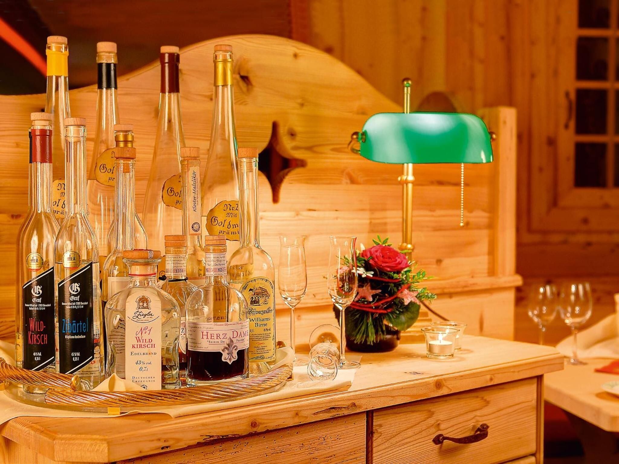 Una barra de madera con varias botellas de licor, una lámpara verde y un pequeño arreglo floral.