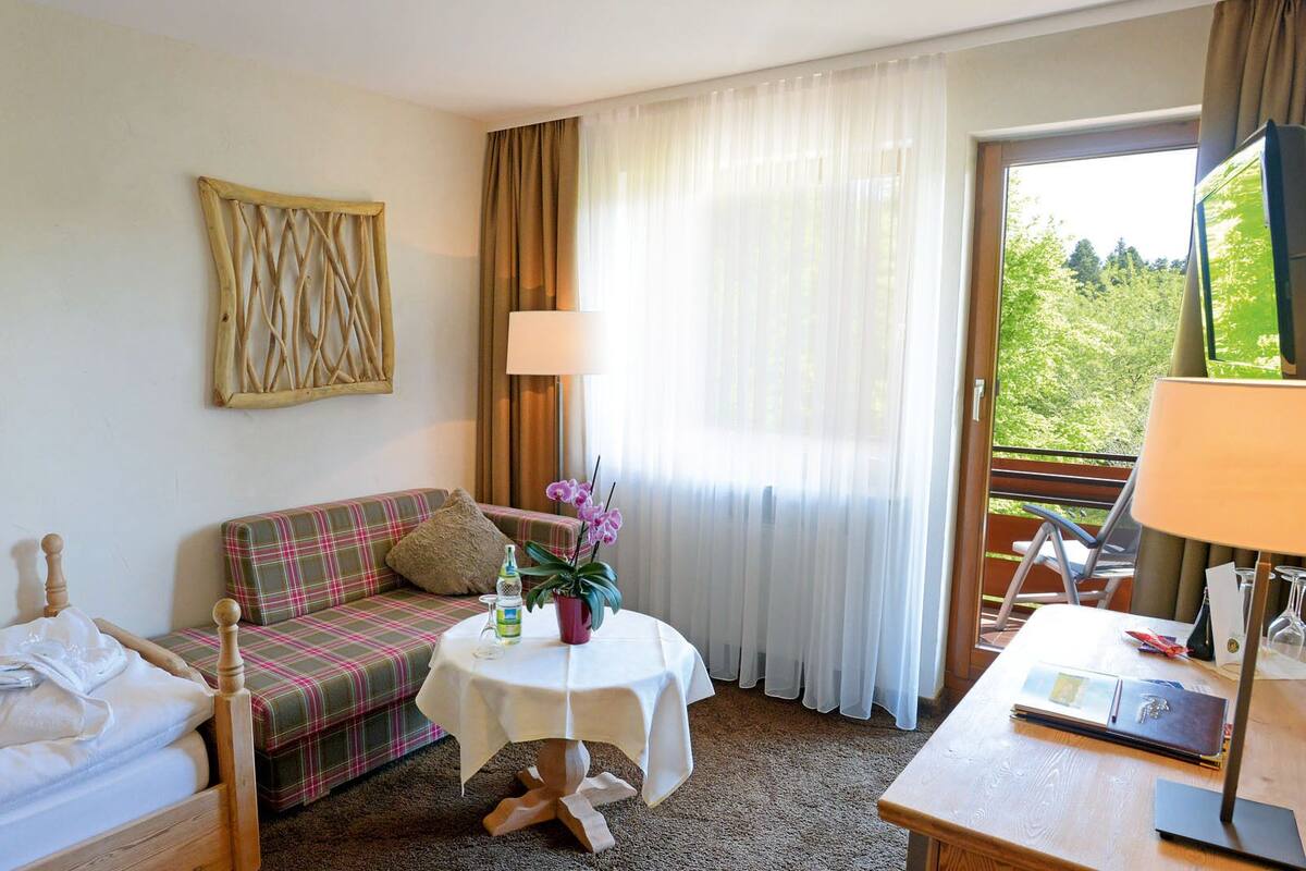 A hotel room with a bed, a sofa, a small table with a lamp, and a view of greenery through the window.