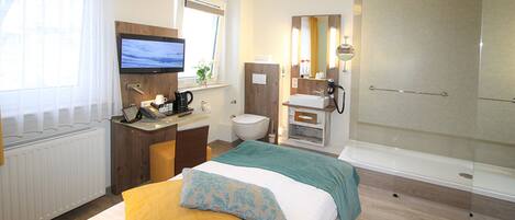 A hotel room with a bed, a desk with a computer, a TV, a bathroom with a shower, and a window with curtains.
