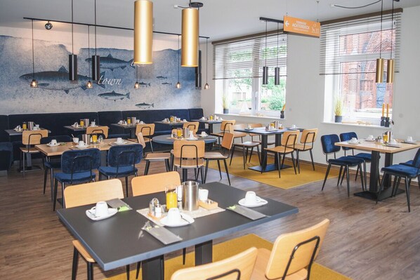 Un restaurante moderno con varias mesas y sillas, un mural de peces en la pared y ventanas amplias con persianas.