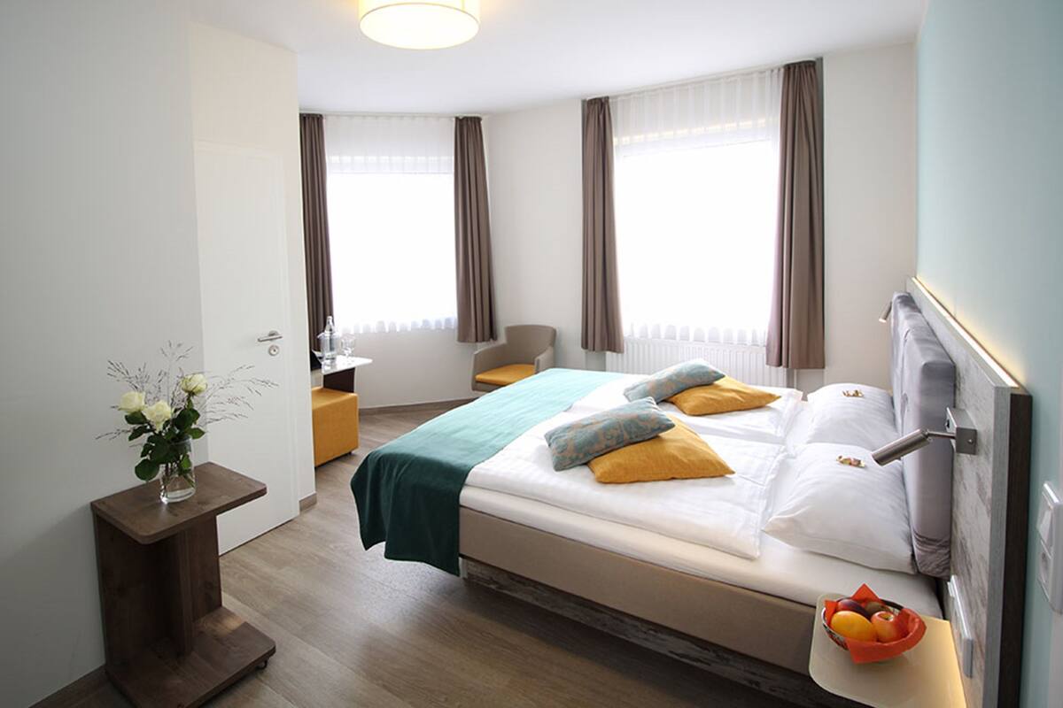 A modern hotel room with a large bed, a bedside table with a vase of flowers, a chair, and a fruit bowl.