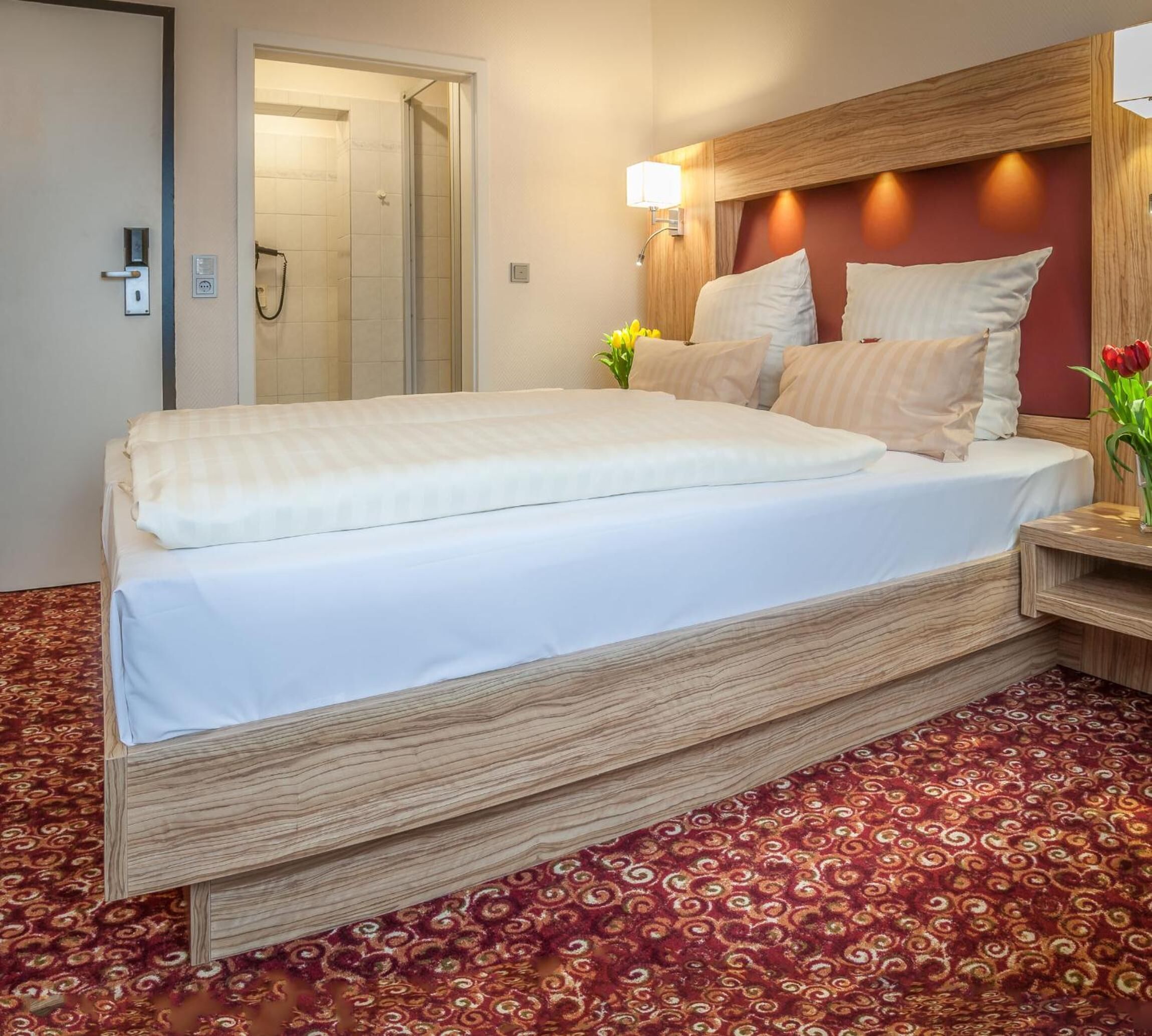 A hotel room with a large bed, a wooden headboard, a bedside table with a lamp, and a patterned carpet.