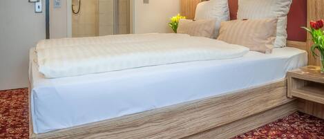 Una habitación de hotel con una cama grande, cabecera de madera, mesita de noche con lámpara y alfombra estampada.