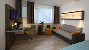Una habitación de hotel moderna con un sofá, un escritorio con una computadora, una silla, una mesita con un florero y una ventana con cortinas.