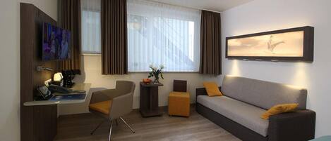 A modern hotel room with a sofa, a desk with a computer, a chair, a small table with a vase, and a window with curtains.
