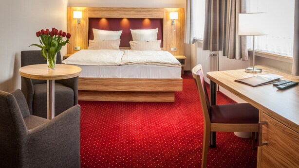 Habitación de hotel con una cama, un escritorio, una silla, una mesita con un jarrón de flores y una alfombra roja.