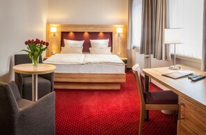 Een hotelkamer met een bed, een bureau, een stoel, een kleine tafel met een bloevas en een rood tapijt.