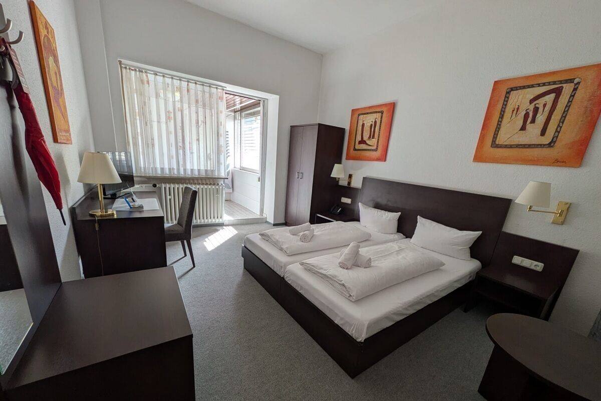 A hotel room with a large bed, a desk, a chair, and two paintings on the wall.