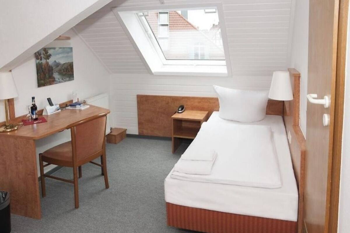 A compact room with a bed, a desk, a chair, a small table, a lamp, and a window.