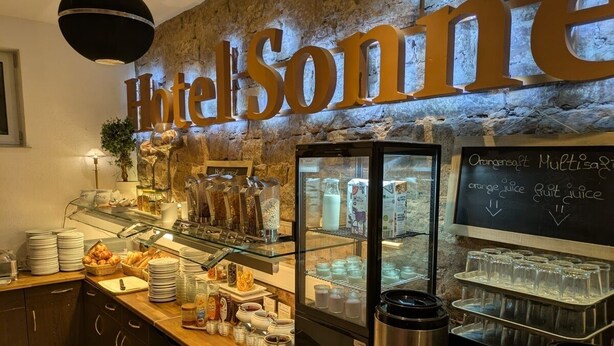 A hotel breakfast area with a stone wall, a large sign reading 'Hotel Sonine,' a glass display case with food items, and a chalkboard menu. - Hotel Sonne (Karlsruhe)